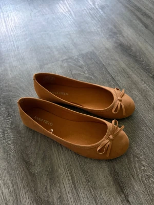 Bruna ballerinaskor med rosett - Snygga ballerinaskor från Anna Field i en varm brun färg. Skorna har rund tå och en söt rosett framtill. Tillverkade i ett mjukt material med matt finish som ger en stilren look. Perfekta att matcha med jeans eller klänning.
