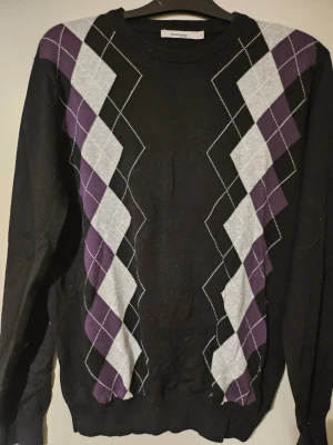 Svart argyle-stickad tröja Dressmann - Stickad tröja från Dressmann i svart med klassiskt argyle-mönster i lila och grått på framsidan. Tröjan har rund halsringning och långa ärmar. Perfekt för dig som gillar retrovibbar och vill ha något stilrent men ändå med lite edge.
