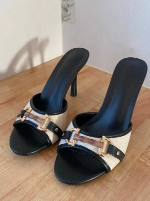 Svarta och beige klackar - Säljer ett par stilrena sandaler med hög klack i svart och beige. Ovandelen är i textil med svarta läderdetaljer och guldfärgade metalldekorationer samt en brun detalj framtill. Öppen tå och elegant. Skit snygga men måste säljas eftersom har för många! Köpt från Tunisien! 