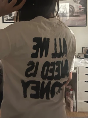 Vit t-shirt med svart textprint - Vit t-shirt med svart, stor text på ryggen där det står 'ALL WE NEED IS MONEY'. T-shirten har en avslappnad passform och korta ärmar. Perfekt för dig som gillar statement-plagg och enkel stil.