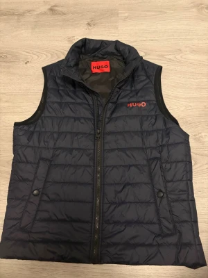 Mörkblå dunväst från HUGO boss - Snygg mörkblå dunväst från HUGO boss med diskret quiltad design och röd logga på bröstet. Västen har hög krage, dragkedja framtill och två fickor med tryckknappar. Perfekt för lager-på-lager och streetstyle.