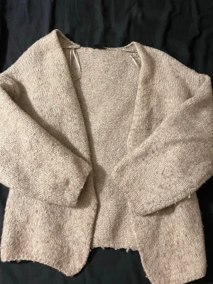 Beige stickad öppen kofta - Mysig beige stickad kofta med öppen front och lång ärm. Koftan har en avslappnad passform och är tillverkad i ett mjukt, grovt stickat material som passar perfekt för lager på lager. Enkel och stilren design utan knappar eller dragkedja.