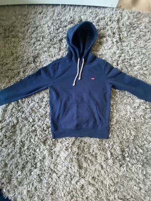 Marinblå hoodie från Levi's XS - Säljer en marinblå hoodie från Levi's i storlek XS. Tröjan har huva med vita snören, en klassisk magficka och långärmade ärmar. Diskret Levi's-logga på bröstet. Perfekt för chill dagar och enkel att matcha med jeans eller joggers.