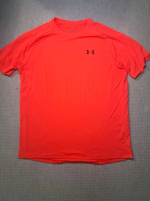 Neonröd tränings-tshirt Under Armour - Snygg neonröd tränings-tshirt från Under Armour med korta ärmar och rund hals. Tillverkad i ett lätt och snabbtorkande funktionsmaterial som passar perfekt till gymmet eller löprundan. Diskret logga på bröstet och nacken.