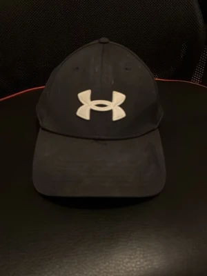 Svart keps från Under Armour - Snygg svart keps från Under Armour med böjd skärm och stort vitt logotryck framtill. Klassisk sexpanel-design och justerbar passform bak. Perfekt för en sportig och avslappnad stil.