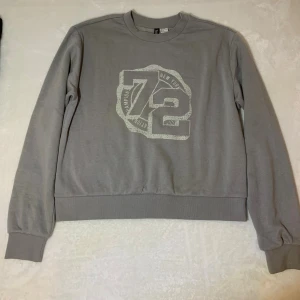 Grå sweatshirt med tryck från H&M - Grå sweatshirt från H&M med stort vitt tryck på bröstet där det står '72' och text som 'New York' och 'Champion'. Tröjan har rund halsringning, ribbade muddar och är tillverkad i mjukt bomullsmaterial. Perfekt för en avslappnad streetwear-look.