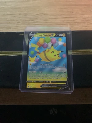 Pokémonkort Flying Pikachu V - Säljer ett Pokémonkort Flying Pikachu V med färgglada ballonger och Pikachu i fokus. Kortet har en blå bakgrund och är innesluten i ett genomskinligt plastfodral. Perfekt för samlare eller Pokémonfans som vill utöka sin kortsamling.