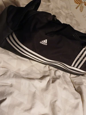 Svart sportbag från Adidas - Snygg svart sportbag från Adidas med klassiska vita ränder och logga på sidan. Väskan har axelrem, bärhandtag och flera fack med dragkedja. Perfekt för gymmet eller träningen. Tillverkad i slitstarkt syntetmaterial.