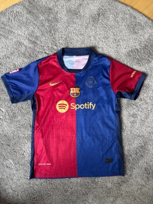 FC Barcelona matchtröja Nike - Säljer en officiell FC Barcelona matchtröja från Nike i rött och blått med klubbmärke, Spotify-logga och guldfärgad Nike-swoosh på bröstet. Tröjan har korta ärmar, rund hals och är tillverkad i ett lätt, ventilerande funktionsmaterial.