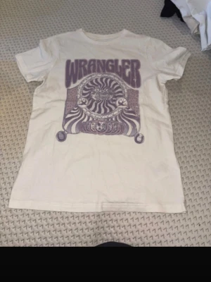 Vit Wrangler t-shirt med tryck - Cool vit t-shirt från Wrangler med stort retroinspirerat tryck i lila på framsidan. Trycket har ett psykedeliskt mönster med sol och måne samt zebraränder. Klassisk passform och rund hals. Perfekt för dig som gillar vintage vibes.