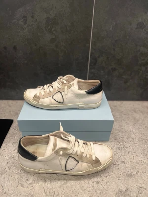 Vita sneakers från Philippe Model - Snygga vita sneakers från Philippe Model med svarta detaljer på hälen och den ikoniska sköldloggan på sidan. Skorna har klassisk låg profil, vita snören och ovandel i läder. Perfekta för en avslappnad streetstyle-look. Storlek 44. Hör av dig för mer bilder eller funderingar ! / Isak