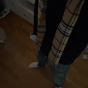 Beige rutig halsduk - Klassisk beige halsduk från Burberry med det ikoniska rutmönstret i svart, vitt och rött. Halsduken har fransar i ändarna och är tillverkad i ett mjukt material som känns lyxigt och stilrent. Perfekt accessoar för att lyfta din outfit.