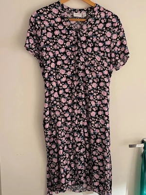 Blommig kortklänning från Vila - Supersöt kortklänning från Vila med svart botten och blommigt mönster i rosa och ljusblått. Klänningen har korta ärmar, rund halsringning och en avslappnad passform. Perfekt för vår och sommar! Knytband i midjan. Använd 1 gång.