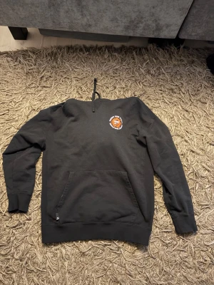 Svart hoodie från Puma med tryck - Svart hoodie från Puma med huva och känguruficka. Framtill finns ett litet färgglatt tryck på bröstet och på ryggen flera grafiska tryck i vitt, lila och orange med text och motiv. Perfekt för en avslappnad streetwear-look.