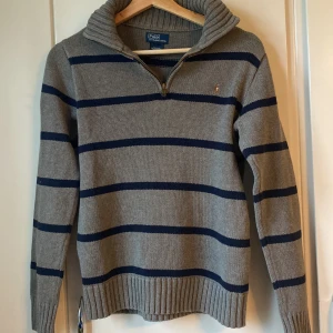 Half Zip Ralph Lauren - Snygg stickad äkta tröja från Polo Ralph Lauren i grått med marinblå ränder. Tröjan har krage, half zip-dragkedja och klassisk broderad logga på bröstet. långa ärmar med ribbade muddar.