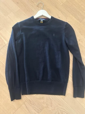 Ralph Lauren Tröja - Klassisk mörkblå stickad tröja från Polo Ralph Lauren med rund halsringning och diskret broderad logga på bröstet. Tröjan har långa ärmar och ribbade muddar vid ärmslut och nederkant. Perfekt till jeans eller chinos.
