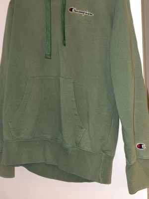 Grön hoodie från Champion - Snygg grön hoodie från Champion med broderad logga på bröstet och ärmen. Klassisk känguruficka framtill och justerbar huva med matchande snören. Perfekt för en avslappnad och sportig stil. Mjuk bomullsmix som känns skön mot huden. Lite knopprig och några fläckar men dem syns inte 