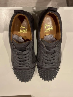 Svarta Louboutin sneakers med nitar - Säljer ett par svarta sneakers från Christian Louboutin med mattsvarta nitar på tån och klassisk röd sula. Skorna har snörning, är låga och har en lyxig känsla med läderdetaljer och ikonisk logga på innersulan.kommer även i låda skorhorn och påsar till även kvitto