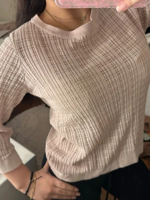 Ljusrosa ribbad tröja med struktur - Säljer en ljustosatröja med ribbad struktur och rund halsringning. Tröjan har långa ärmar och är tillverkad i ett mjukt, bekvämt material. Perfekt för en stilren och avslappnad look.