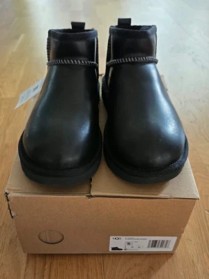 Svarta stövletter från UGG - Snygga svarta stövletter från UGG i slätt läder med kontrastsömmar och låg ankelhöjd. Skorna har en robust sula och minimalistisk design som passar till många olika stilar. Klassisk UGG-logga på sidan och bekväm passform.