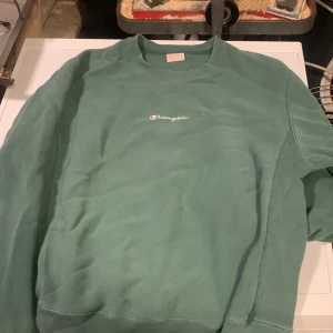 Grön sweatshirt från Champion - Snygg grön sweatshirt från Champion med broderad logga på bröstet. Klassisk rund halsringning och ribbade muddar vid ärmslut och nederkant. Perfekt för en avslappnad och sportig stil. Liten skada på bröstet, se bild! Skulle säga att den passar M också. 