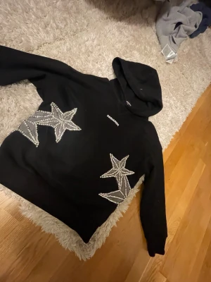 Svart hoodie med stjärnor från Dsquared2 - Knappt använd Snygg svart hoodie från Dsquared2 med huva och blingade stjärnor i silver på framsidan. Märkesloggan sitter diskret på bröstet. Hoodien har en avslappnad passform och är tillverkad i mjukt bomullsmaterial. Perfekt för dig som vill sticka ut med coola detaljer.