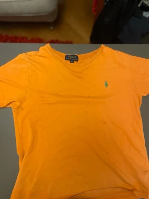 Orange t-shirt från Polo Ralph Lauren (fläckig) - Den är tyvärr fläckig därpå priset Snygg orange t-shirt från Polo Ralph Lauren med klassisk rund hals och det ikoniska gröna logobroderiet på bröstet. Tillverkad i mjuk bomull och har en normal passform. Perfekt för dig som vill sticka ut med färg.