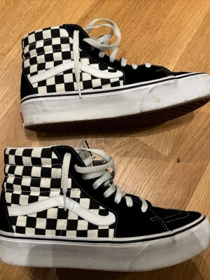 Vans svartvita checkerboard high tops stl  40,5 - Säljer ett par klassiska Vans sneakers med svartvitt checkerboard-mönster och vita detaljer. Skorna är high tops med vit sula, vita skosnören och den ikoniska Vans-sidan i vitt. Ovandelen är i canvas och mocka för en skön och cool look.