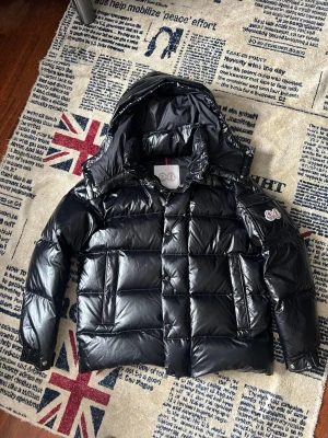 Svart dunjacka från Moncler - Snygg svart dunjacka från Moncler med glansig finish och stor huva. Jackan har två stora framfickor med lock och tryckknapp samt Moncler-logga på ärmen. Perfekt för kalla vinterdagar och riktigt trendig look. NFC