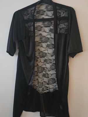 Svart negless med spetsdetaljer - Snygg svart negless ii satinliknande material med genomskinliga spetsdetaljer på rygg och axlar. Halvlånga ärmar och öppen framtill, perfekt för en chill dag hemma. Spetsen har blommönster som ger en extra elegant touch.           Storlek: XS/S Mått: Axel till axel: 38 cm Ärm: 30 cm Nacke till fåll: (längd rygg): 87 cm   Tänk på att måtten är så exakta jag kan mäta men kan avvika, mäter givetvis dem när jag lägger in annonsen.  Skick: NY  SAMFRAKTAR GÄRNA!  Köp det du gillar och om det blir mer 