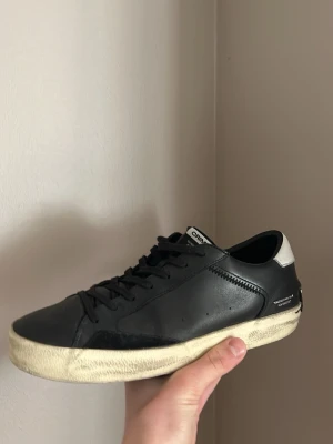 Svarta Crime london sneakers i läder - Stilrena svarta sneakers från Crimblé i slätt läder med vit kontrastdetalj på hälen och textdetaljer. Skon har svarta skosnören och en vit, något smutsad sula för en cool, använd look. Perfekta för dig som gillar minimalistisk och modern stil.