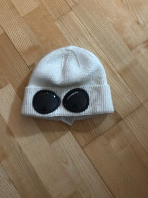  C.P. Company mössa - Vit ribbstickad mössa från C.P. Company med ikoniska svarta goggles framtill. Tillverkad i 100% bomull och har en uppvikt kant. Perfekt accessoar för dig som vill sticka ut och addera streetwear-vibe till din outfit.