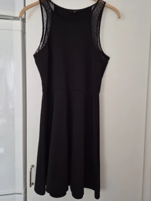 Klänning - Snygg svart klänning från H&M med meshdetaljer vid axlarna och ärmlöst snitt. Klänningen har en rund halsringning och lätt utställd kjol som ger en skön passform. Perfekt för dig som gillar enkel men stilren design.