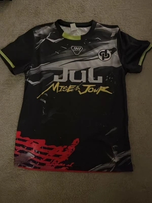 Svart sporttröja JUL Mise à Jour - Snygg svart sporttröja från JUL med trycket 'Mise à Jour' i gult och vitt framtill. Tröjan har korta ärmar, rund hals och detaljer i rött, vitt och limegrönt. Tillverkad i ett lätt och snabbtorkande material, perfekt för träning eller sport.