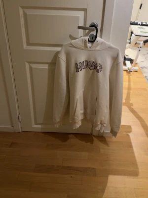 Vit hoodie från Hugo med tryck - Säljer en vit hoodie från Hugo med coolt tryck på bröstet. Hoodien har luva, ribbade muddar och en klassisk känguruficka framtill. Perfekt för dig som gillar enkel men snygg streetwear.