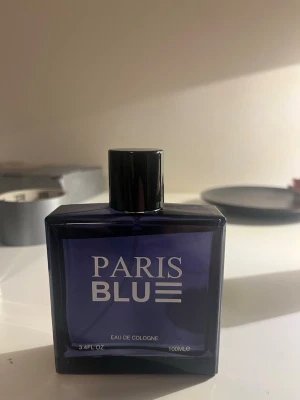 Paris Blue Eau de Cologne 100ml - Paris Blue Eau de Cologne i en stilren, mörkblå glasflaska med svart lock. Flaskan har en fyrkantig form och rymmer 100 ml. Texten på framsidan är vit och designen känns modern och enkel.