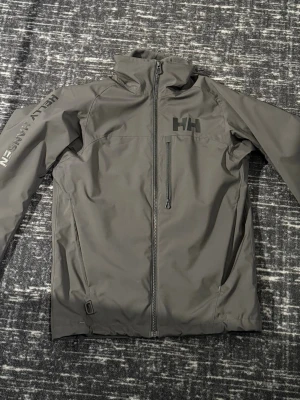 Helly Hansen jacka - Helly Hansen Lifalot shelljacka med hög krage och dragkedja framtill. Jackan har en bröstficka med dragkedja och logga på bröstet samt ärmen. Tillverkad i ett slitstarkt, vattenavvisande material som passar perfekt för blåsiga dagar.