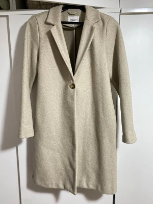 Beige kappa från ONLY - Stilren beige rock från ONLY med klassisk krage och enkelknäppning. Rak passform och långa ärmar ger en clean look. Original pris 600kr