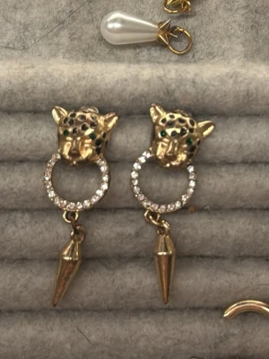 Guldörhängen leopard med strass - Unika örhängen i guldfärg med leopardhuvud och gröna detaljer för ögonen. Ringen under huvudet är dekorerad med gnistrande strass och avslutas med en spetsig hängande detalj. Djurinspirerad design som sticker ut.