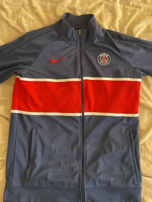 PSG träningsjacka från Nike blå/röd - Snygg PSG träningsjacka från Nike i blått med röd och vit rand över bröstet. Jackan har dragkedja, hög krage och två fickor med dragkedja. PSG-logga och Nike Swoosh på bröstet. Perfekt för fotbollsträning eller streetwear. Dragkedjaan är dock lite trög men fungerar! 