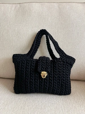 Svart virkad handväska med gulddetalj - Unik svart handväska i virkat material med två handtag och snygg guldfärgad vridlåsdetalj framtill. Väskan har en mjuk, rektangulär form och är perfekt för dig som gillar en personlig och trendig accessoar. Passar till alla outfits och ger en cool touch.