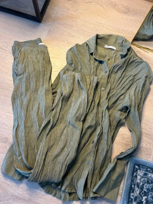 Olivgrönt plisserat loungewear-set - Snyggt loungewear-set från Bershka i olivgrönt plisserat tyg. Setet består av en långärmad skjorta med knappar och matchande byxor med avslappnad passform. Perfekt för chill dagar hemma eller när du vill vara bekväm men ändå se stylad ut.