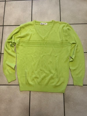 Limegrön stickad tröja storlek M/L - Stickad limegrön tröja med V-ringning och diskreta ränder över bröstet. Tröjan har långa ärmar och ribbade muddar vid ärmslut och nederkant. Perfekt för dig som gillar färg och vill sticka ut lite extra.