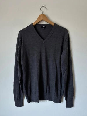 Mörkgrå V-ringad tröja från Uniqlo - Stilren mörkgrå stickad tröja från Uniqlo i 100% ull med V-ringning och långa ärmar. Tillverkad i tunt, mjukt material som passar perfekt för lager på lager. Klassisk passform och ribbade muddar vid ärmslut och nederkant. 8D21