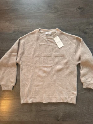 Beige stickad tröja från Lis Bonne - Beige stickad tröja från Lis Bonne med rund halsringning och ribbade muddar. Tröjan har ett diskret, upphöjt mönster framtill som ger en cool detalj. Perfekt för dig som gillar stilrena och mjuka plagg i neutrala färger.