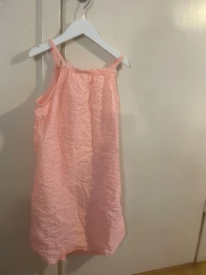 Rosa randig sommarklänning - Söt kort klänning i ljusrosa med tunna vita ränder. Klänningen har smala axelband och är tillverkad i ett mjukt, luftigt bomullstyg som passar perfekt för varma dagar. Enkel och avslappnad modell med ledig passform.