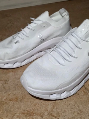 Vita On Running sneakers - Säljer ett par helvita sneakers från On Running med låg profil och modern design. Skorna har ventilerande meshmaterial, elastiska snören och en chunky sula med On's karakteristiska CloudTec-dämpning. Perfekta för dig som gillar stilrena och sportiga skor. SNABB AFFÄR 500KR