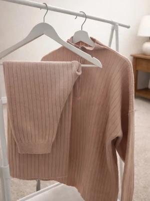Beige ribbat loungewear-set - Mysigt loungewear-set i beige med ribbad struktur. Setet består av en långärmad tröja med halv dragkedja och matchande byxor med resår i midjan och benslut. Passar S/M