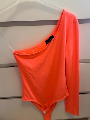 Neonorange oneshoulder topp - Säljer en neonorange oneshoulder topp från PrettyLittleThing. Toppen har lång ärm på ena sidan och är axelbandslös på andra, vilket ger en cool asymmetrisk look. Materialet känns mjukt och stretchigt, perfekt för att sticka ut. Oanvänd 