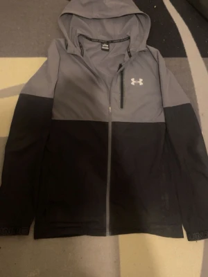 Grå och svart vindjacka Under Armour - Snygg tvåfärgad vindjacka från Under Armour i grått och svart. Jackan har huva, hel dragkedja framtill och en bröstficka med dragkedja. Tillverkad i lätt och slitstarkt material, perfekt för blåsiga dagar. Under Armour-logga på bröstet.